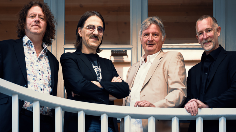 Vier Männer in Anzügen stehen hinter einem weißen Geländer und lächeln in die Kamera. Hintergrund aus Holz und Glas., © Foto Privat Vier Männer in Anzügen stehen hinter einem weißen Geländer und lächeln in die Kamera. Hintergrund aus Holz und Glas., © Foto Privat