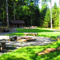 Ein Grillplatz im Wald mit Holztischen, Bänken und einer Feuerstelle, umgeben von hohen Bäumen und grünem Gras., © Locher Fotodesign & Manufaktur