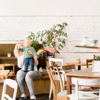Gemütliches Café mit spielenden Kindern und Erwachsenen. Eine Bedienung serviert Getränke. Pflanzen und Holzmöbel schaffen eine einladende Atmosphäre., © Sven Weber Gemütliches Café mit spielenden Kindern und Erwachsenen. Eine Bedienung serviert Getränke. Pflanzen und Holzmöbel schaffen eine einladende Atmosphäre., © Sven Weber