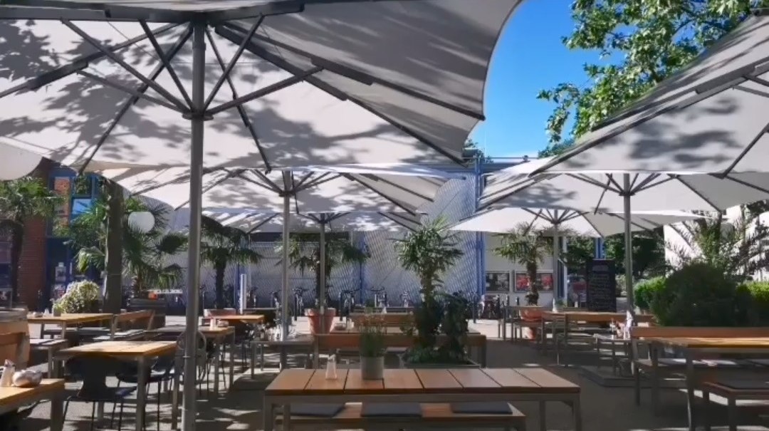 Ein Café-Außenbereich mit großen Sonnenschirmen, Holztischen und Stühlen. Pflanzen und Bäume spenden Schatten an einem sonnigen Tag., © Blauer Engel GmbH