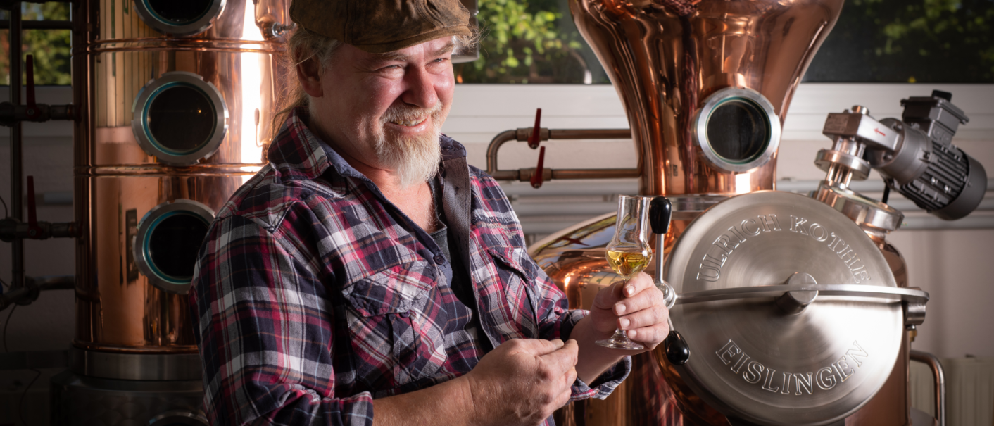 Mann in kariertem Hemd hält ein Glas mit goldenem Getränk vor einer Kupferdestille. Er lächelt und trägt eine Mütze. Im Hintergrund sind Fenster und Bäume sichtbar., © Brau- & Brennwerkstatt