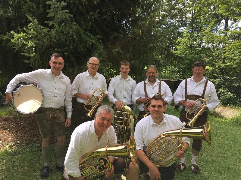 Eine Blaskapelle in traditioneller Kleidung posiert mit Instrumenten im Freien vor Bäumen. Die Musiker tragen weiße Hemden und Lederhosen., © Alb 7 Eine Blaskapelle in traditioneller Kleidung posiert mit Instrumenten im Freien vor Bäumen. Die Musiker tragen weiße Hemden und Lederhosen., © Alb 7