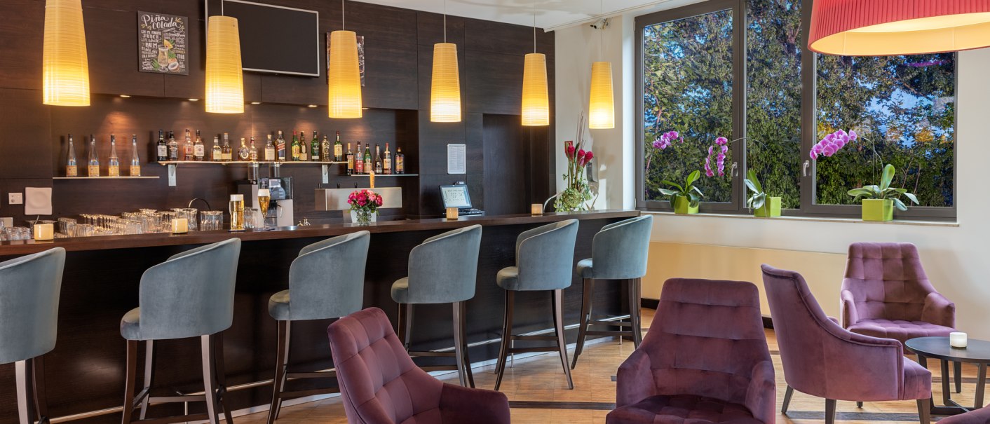 Elegante Bar mit lila Sesseln, Barhockern und hängenden Lampen. Große Fenster mit Blick auf Bäume und Orchideen., © NH Hotel Stuttgart Airport
