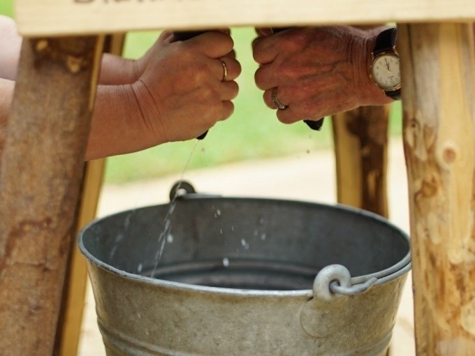 Zwei Personen melken eine Kuhattrappe, während Wasser in einen Metalleimer tropft. Eine Person trägt eine Uhr., © Cool-Tours StattReisen Zwei Personen melken eine Kuhattrappe, während Wasser in einen Metalleimer tropft. Eine Person trägt eine Uhr., © Cool-Tours StattReisen