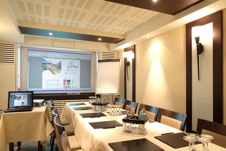 Moderner Konferenzraum mit Tisch, Stühlen, Laptop, Projektor und Flipchart. Getränke und Schreibmaterialien sind bereitgestellt., © Römerhof Hotelbetriebs GmbH