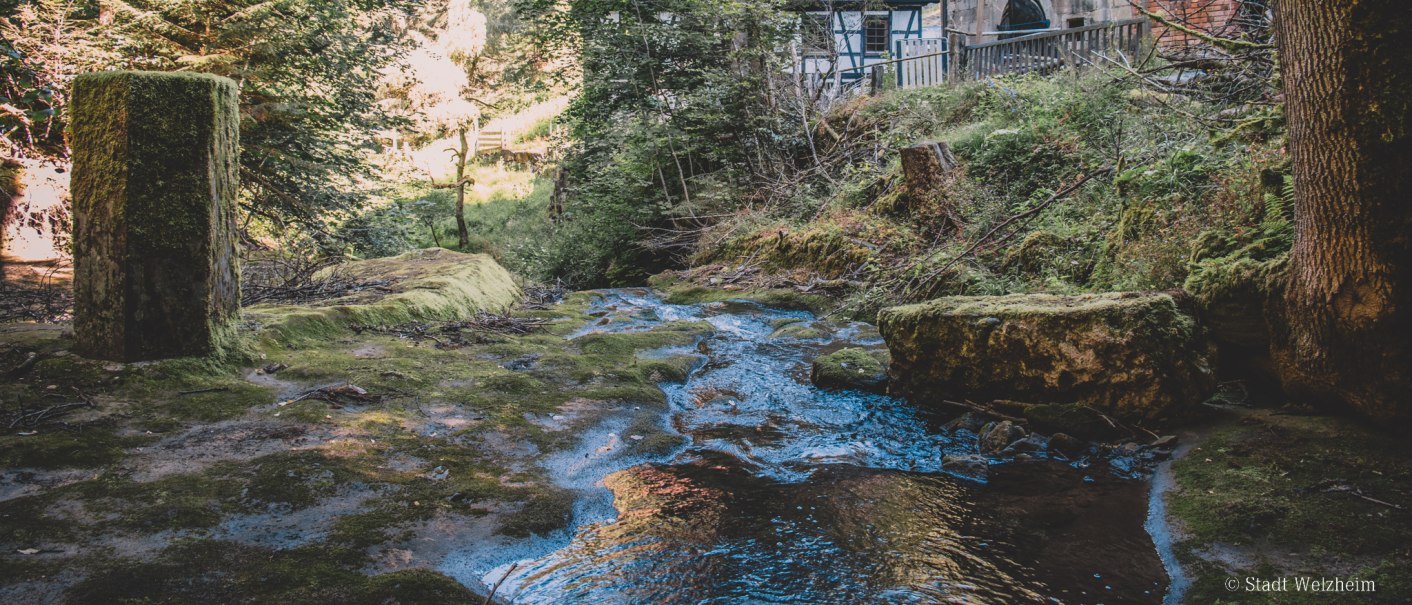 Ein Bach fließt durch einen Wald, vorbei an einem alten Fachwerkhaus. Moosbewachsene Steine und Bäume umgeben die Szene., © Stadt Welzheim