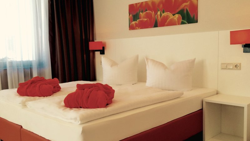 Ein gemütliches Hotelzimmer mit Doppelbett, roten Bademänteln und einem Tulpenbild an der Wand. Die Beleuchtung ist warm und einladend., © TOMAS Ein gemütliches Hotelzimmer mit Doppelbett, roten Bademänteln und einem Tulpenbild an der Wand. Die Beleuchtung ist warm und einladend., © TOMAS