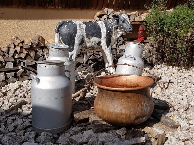 Eine dekorative Szene mit Milchkannen, einem Kupferkessel und einer Kuhfigur auf einem Kiesbett vor einem Holzstapel., © SwabianTravel Eine dekorative Szene mit Milchkannen, einem Kupferkessel und einer Kuhfigur auf einem Kiesbett vor einem Holzstapel., © SwabianTravel