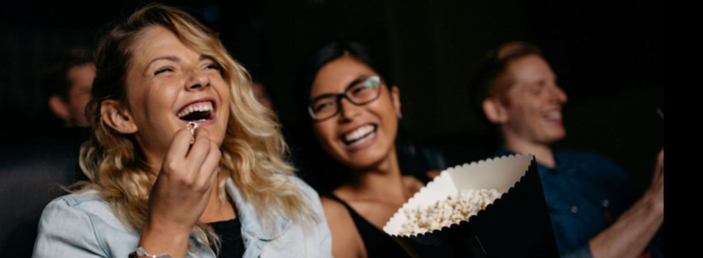 Drei Personen lachen und genießen Popcorn in einem Kino. Die Stimmung ist fröhlich und entspannt.