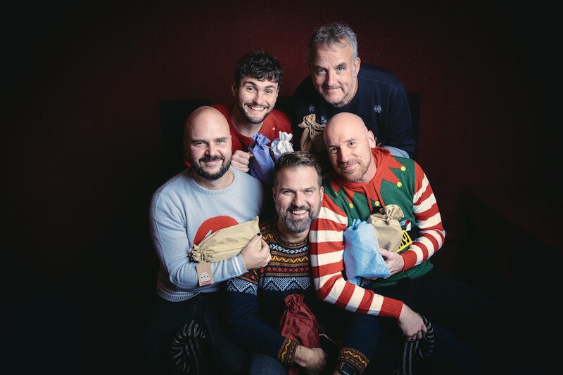 F&uuml;nf M&auml;nner in bunten Weihnachtspullovern halten Geschenkt&uuml;ten und l&auml;cheln fr&ouml;hlich in die Kamera. Die Szene wirkt festlich und herzlich., &copy; Scala Kultur Live gGmbH