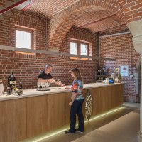 Eine Theke in einer Weinkellerei mit Backsteinwänden. Ein Mann schenkt einer Frau Wein ein. Weinflaschen sind auf der Theke ausgestellt., © Collegium Wirtemberg