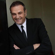 Ein Mann in einem schwarzen Anzug l&auml;chelt und hat die Arme verschr&auml;nkt. Er steht vor einem neutralen Hintergrund., &copy; links im Bild