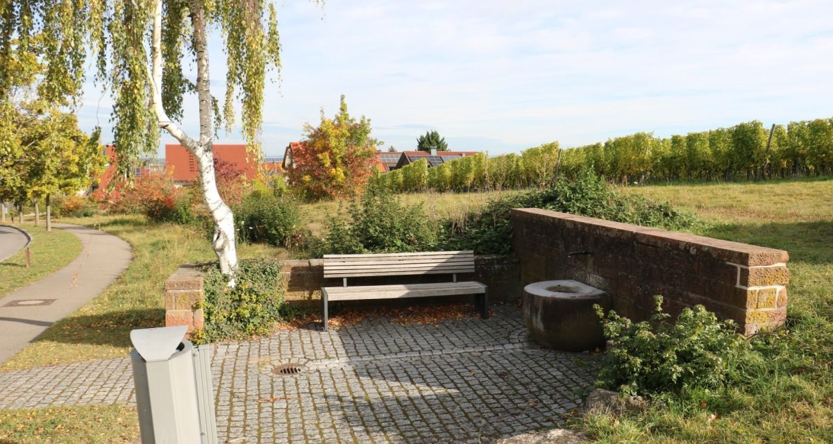 Bank neben einem Baum und Brunnen auf gepflastertem Platz, umgeben von Wiesen und Weinreben. Im Hintergrund sind H&auml;user mit roten D&auml;chern zu sehen., &copy; Remstal Tourismus e.V.