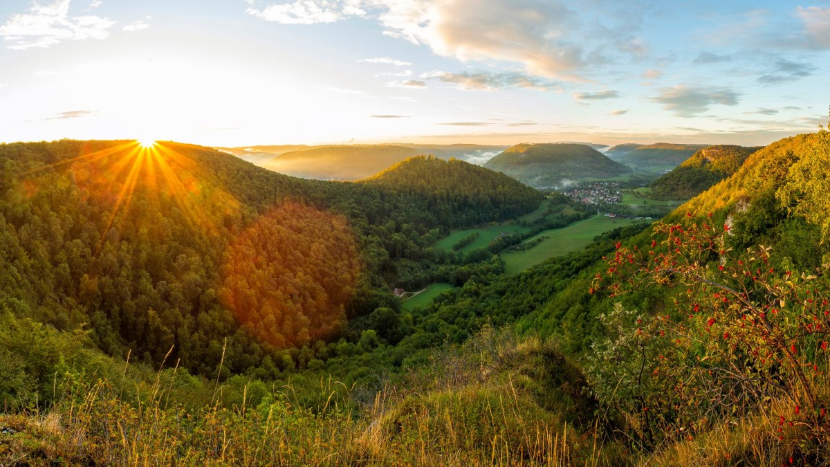 Sonnenaufgang über bewaldeten Hügeln und einem Tal, gesehen vom Badfelsen. Im Vordergrund sind Gräser und Sträucher mit roten Beeren., © Landkreis Göppingen Sonnenaufgang über bewaldeten Hügeln und einem Tal, gesehen vom Badfelsen. Im Vordergrund sind Gräser und Sträucher mit roten Beeren., © Landkreis Göppingen