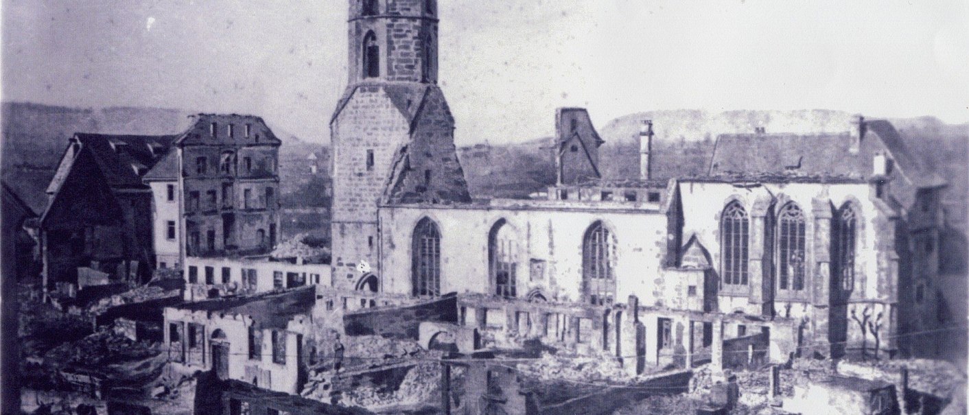Eine Kirche und umliegende Gebäude zeigen schwere Schäden nach dem Stadtbrand von 1868. Ruinen und zerstörte Dächer sind sichtbar., © Kirchenbezirk Schwäbisch Hall-Gaildorf Eine Kirche und umliegende Gebäude zeigen schwere Schäden nach dem Stadtbrand von 1868. Ruinen und zerstörte Dächer sind sichtbar., © Kirchenbezirk Schwäbisch Hall-Gaildorf