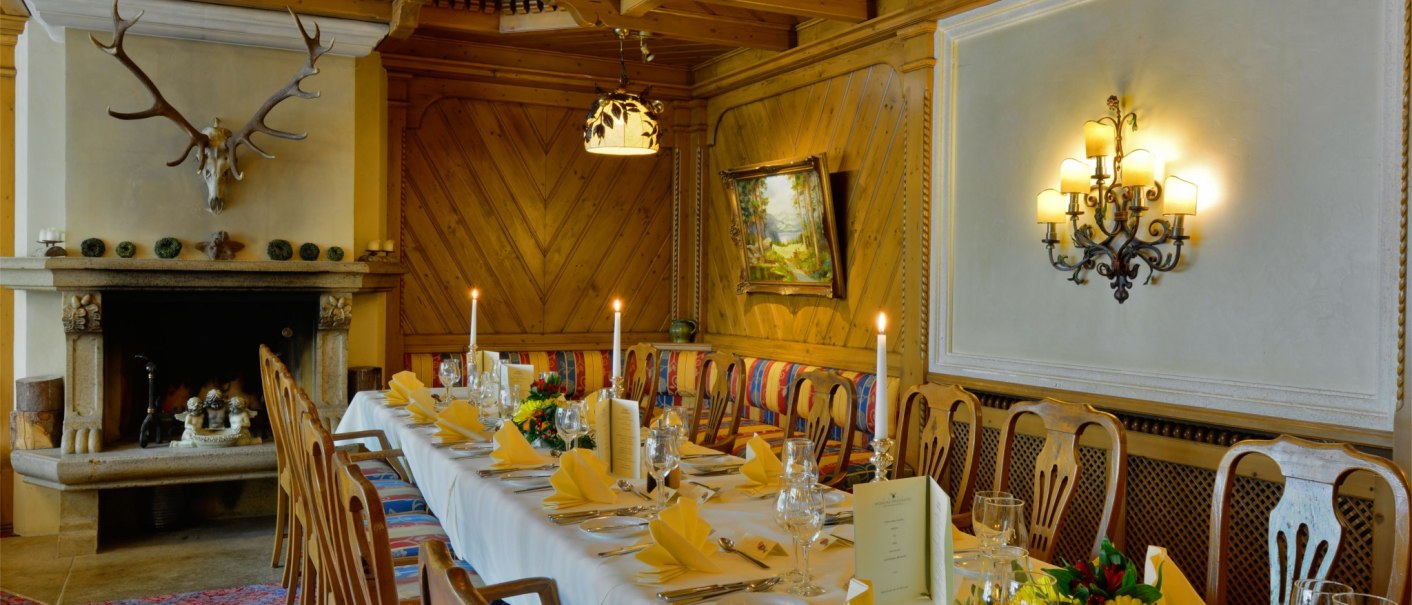 Eleganter Speisesaal mit langem, gedecktem Tisch, Kamin mit Geweih, Holzwänden und Gemälde. Kerzen und Blumenarrangements schmücken den Raum., © TOMAS