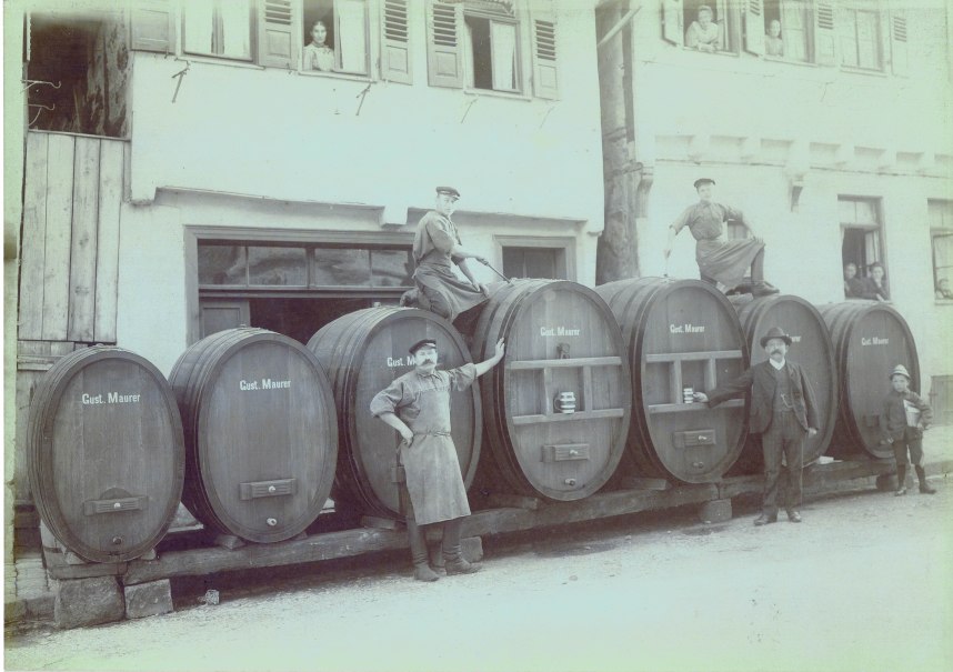 M&auml;nner posieren vor gro&szlig;en Holzf&auml;ssern mit der Aufschrift 'Gust. Maurer'. Personen schauen aus den Fenstern eines Geb&auml;udes., &copy; Stadt Leonberg