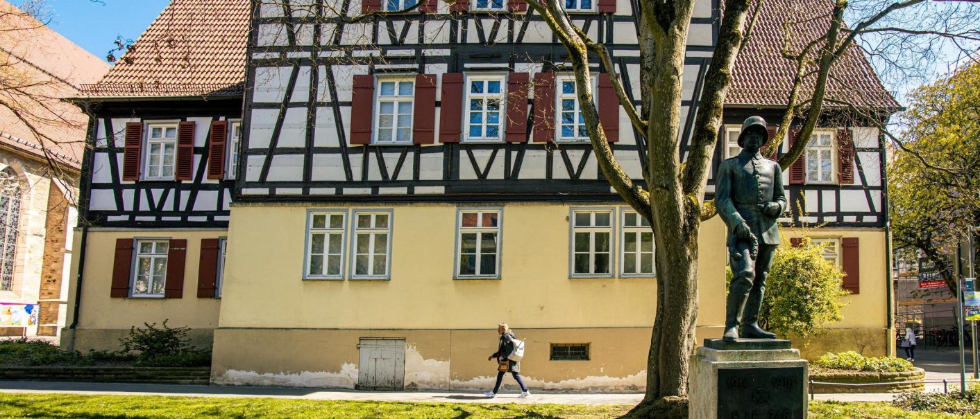 Fachwerkhaus in Kirchheim unter Teck mit einer Statue im Vordergrund. Eine Person geht auf dem Gehweg entlang. Bäume und blauer Himmel im Hintergrund., © Stuttgart-Marketing GmbH, Sarah Schmid