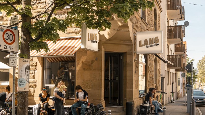 Café Lang an einer Straßenecke mit Menschen, die draußen sitzen. Ein 30er-Zone-Schild und Bäume sind ebenfalls zu sehen., © SMG, Sarah Schmid Café Lang an einer Straßenecke mit Menschen, die draußen sitzen. Ein 30er-Zone-Schild und Bäume sind ebenfalls zu sehen., © SMG, Sarah Schmid