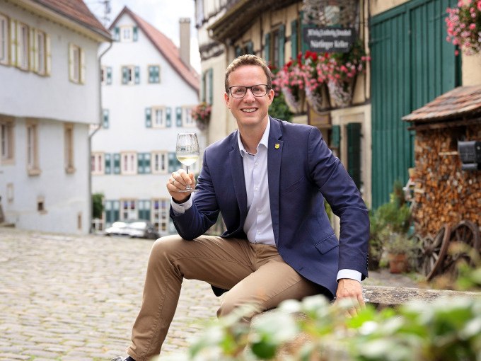 Ein Mann in einem blauen Sakko sitzt in einer malerischen Altstadt und h&auml;lt ein Glas Wein. Im Hintergrund sind Fachwerkh&auml;user und Blumen zu sehen., &copy; Stadt N&uuml;rtingen