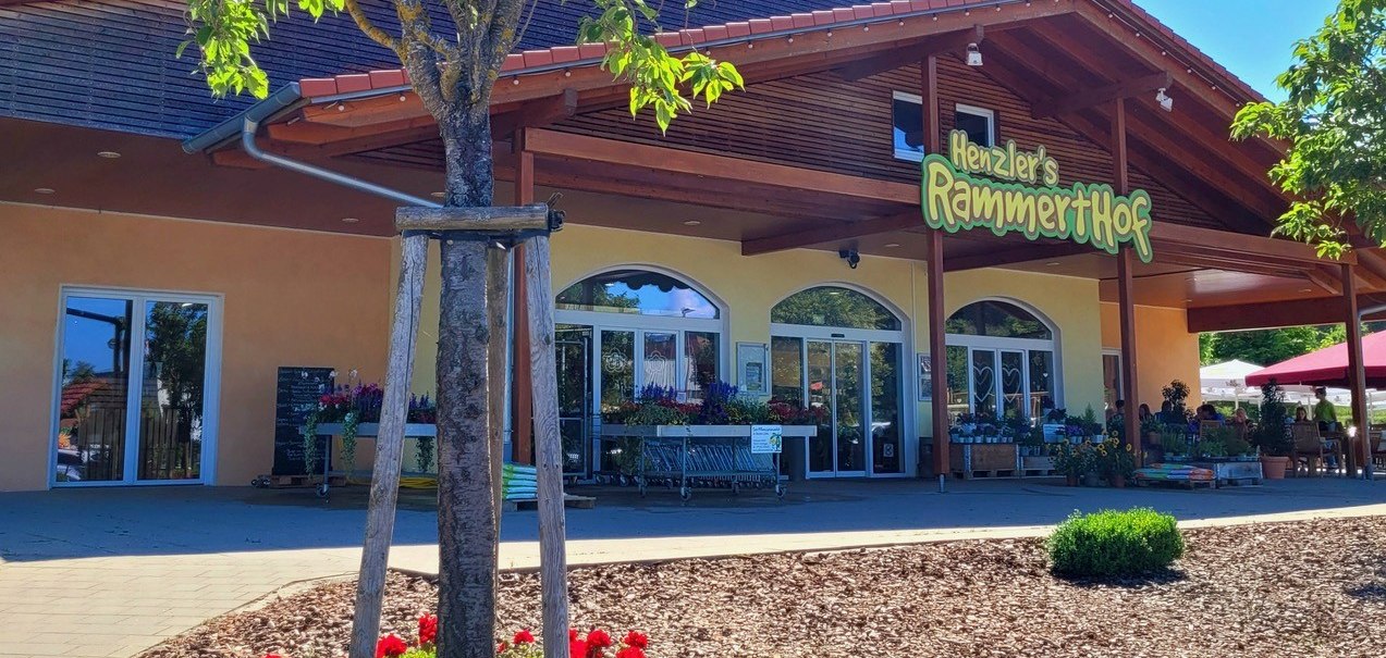 Eingang des Rammerthof-Gebäudes mit Holzverkleidung, Pflanzen und Blumen im Vordergrund. Ein Baum und rote Blumen sind im Vordergrund sichtbar., © Engel/ Rammerthof Eingang des Rammerthof-Gebäudes mit Holzverkleidung, Pflanzen und Blumen im Vordergrund. Ein Baum und rote Blumen sind im Vordergrund sichtbar., © Engel/ Rammerthof