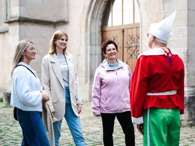 Drei Frauen unterhalten sich mit einer Person in einem bunten Kost&uuml;m vor einem historischen Geb&auml;ude. Die Szene wirkt fr&ouml;hlich und entspannt., &copy; Stadt N&uuml;rtingen