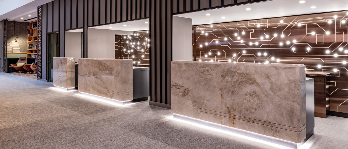 Moderne Rezeption mit Marmor-Theken und beleuchteter Wand. Links gemütliche Sitzecke mit Sesseln und Bücherregal., © Stuttgart Marriott Hotel Sindelfingen