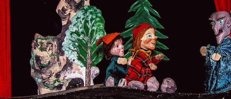 Puppentheater-Szene mit Kasper und anderen Figuren im Wald. Bäume und Felsen im Hintergrund, rote Vorhänge rahmen die Bühne ein., © Prinzessin Gisela Theater