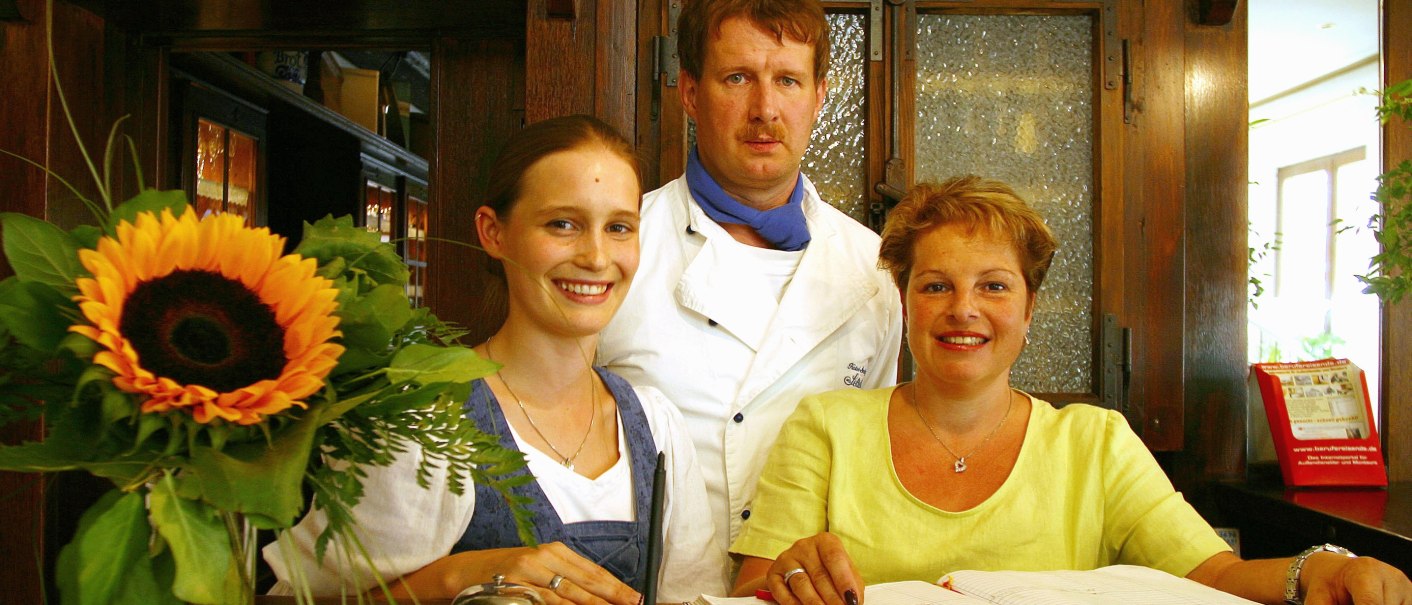 Drei Personen stehen an einer Rezeption: eine Frau in Tracht, ein Mann in Kochkleidung und eine Frau mit einem Buch. Eine Sonnenblume schmückt den Tresen., © R.Storr