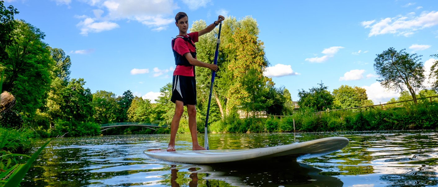 Eine Person steht auf einem Stand Up Paddle Board auf einem Fluss, umgeben von grüner Natur und blauem Himmel., © WTM GmbH Waiblingen