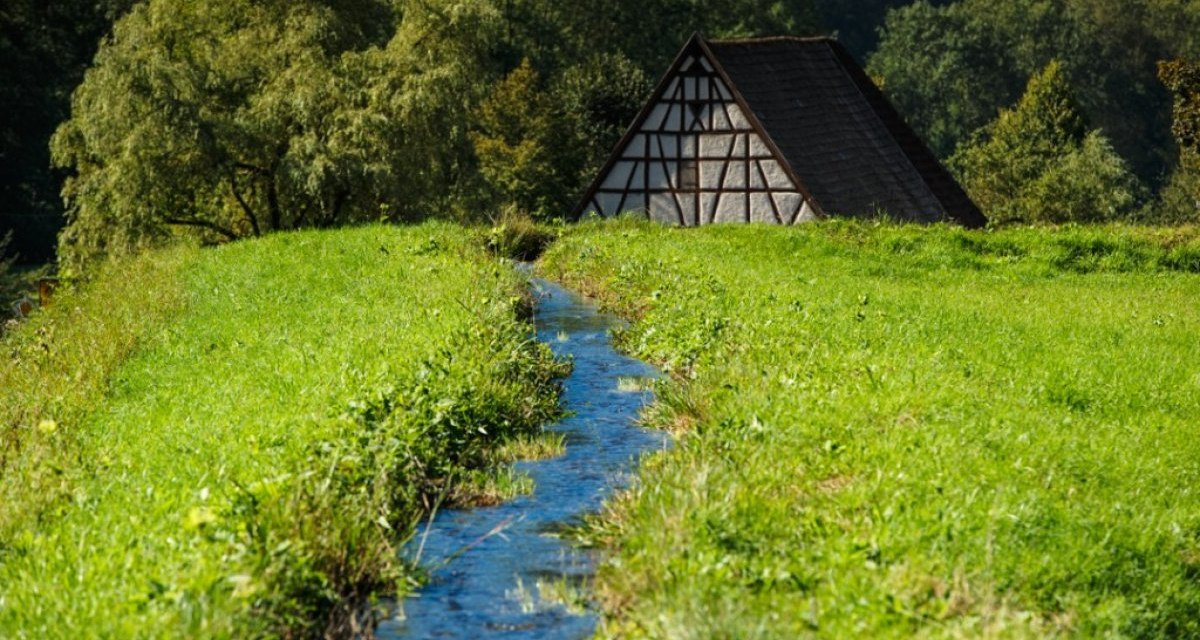 Ein kleines Fachwerkhaus steht im Eybtal, umgeben von grüner Wiese und einem schmalen Bach. Im Hintergrund sind Bäume zu sehen., © Landratsamt Göppingen Ein kleines Fachwerkhaus steht im Eybtal, umgeben von grüner Wiese und einem schmalen Bach. Im Hintergrund sind Bäume zu sehen., © Landratsamt Göppingen