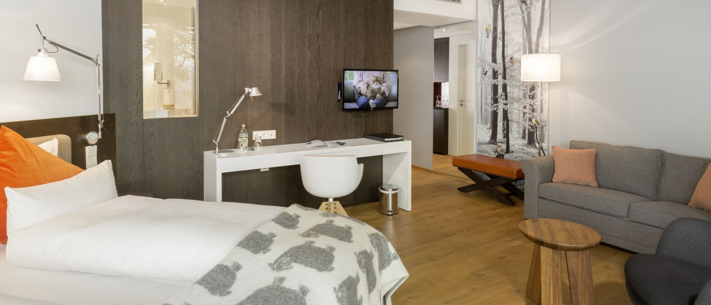 Moderne Hotelsuite mit Bett, Schreibtisch, Sofa und Fernseher. Helle Farben und Holzboden schaffen eine gemütliche Atmosphäre., © Achalm. Hotel GmbH & Co. KG Moderne Hotelsuite mit Bett, Schreibtisch, Sofa und Fernseher. Helle Farben und Holzboden schaffen eine gemütliche Atmosphäre., © Achalm. Hotel GmbH & Co. KG