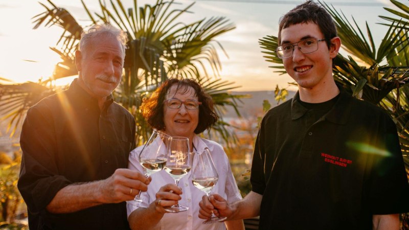 Drei Personen sto&szlig;en mit Weingl&auml;sern im Sonnenuntergang an. Sie stehen vor Palmen und wirken fr&ouml;hlich und entspannt., &copy; Weingut Bayer