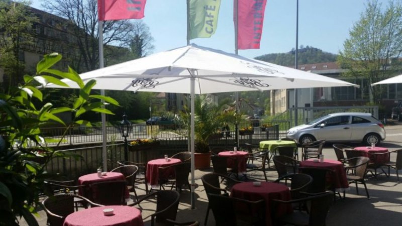 Café-Terrasse mit Tischen, roten Tischdecken und Sonnenschirmen. Im Hintergrund sind eine Straße, Gebäude und grüne Bäume zu sehen., © Intermezzo Café am Bach Café-Terrasse mit Tischen, roten Tischdecken und Sonnenschirmen. Im Hintergrund sind eine Straße, Gebäude und grüne Bäume zu sehen., © Intermezzo Café am Bach