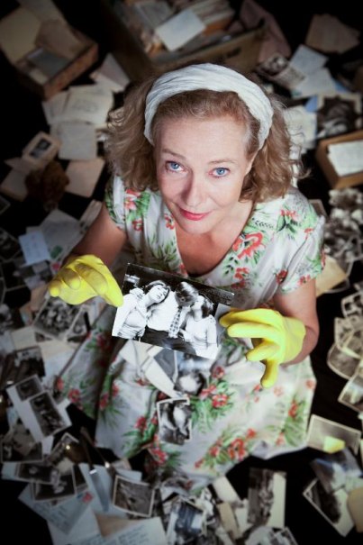 Frau in geblümtem Kleid und gelben Handschuhen hält ein altes Foto, umgeben von verstreuten Fotos und Dokumenten auf dem Boden., © Backnang Frau in geblümtem Kleid und gelben Handschuhen hält ein altes Foto, umgeben von verstreuten Fotos und Dokumenten auf dem Boden., © Backnang