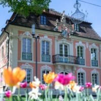 Ein barockes Gebäude mit verzierten Fenstern und Balkon, umgeben von bunten Tulpen im Vordergrund. Ein Kronleuchter hängt im Freien., © Touristik und Marketing GmbH