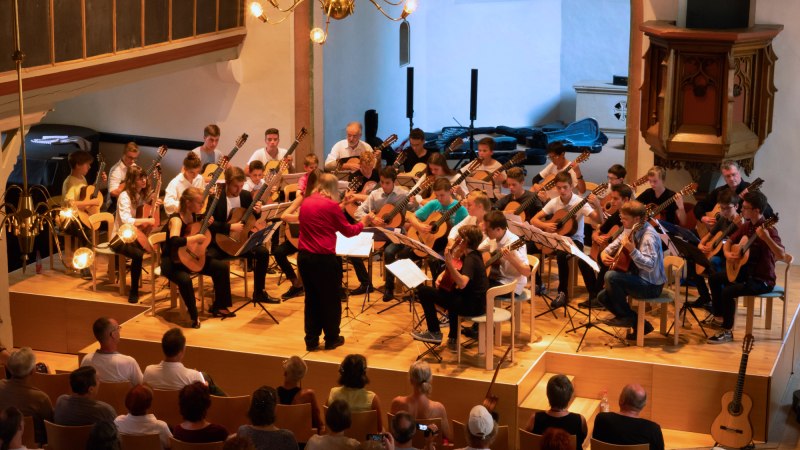 Ein gro&szlig;es Gitarrenensemble spielt in einer Kirche. Der Dirigent steht vorne, w&auml;hrend das Publikum aufmerksam zuh&ouml;rt., &copy; Steffen Brunner