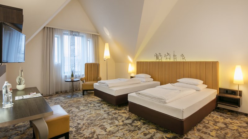 Ein modernes Hotelzimmer mit zwei Einzelbetten, einem Schreibtisch, einem Sessel und einem Flachbildfernseher. Die Einrichtung ist in warmen Tönen gehalten., © ARCOTEL Hotels Ein modernes Hotelzimmer mit zwei Einzelbetten, einem Schreibtisch, einem Sessel und einem Flachbildfernseher. Die Einrichtung ist in warmen Tönen gehalten., © ARCOTEL Hotels