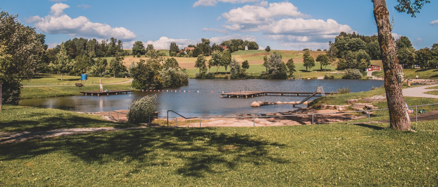 Idyllischer See mit Steg und Rutsche, umgeben von gr&uuml;nen Wiesen und B&auml;umen. Blauer Himmel mit wei&szlig;en Wolken im Hintergrund., &copy; Stadt Welzheim
