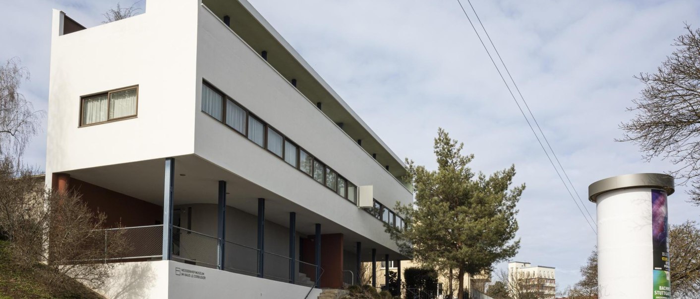 Modernes Gebäude mit weißer Fassade und großen Fenstern, umgeben von Bäumen und einem Plakatständer., © Stuttgart Marketing GmbH, Sarah Schmid