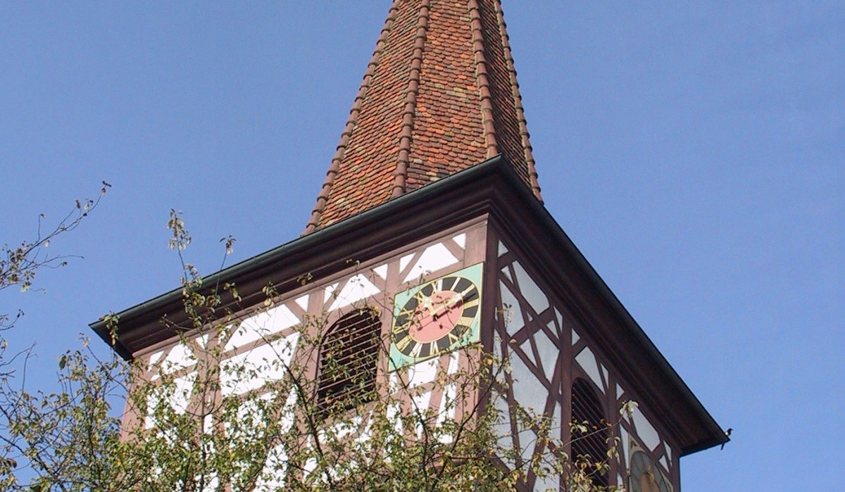 Ein Kirchturm mit Fachwerk und Uhr ragt über Bäume in den blauen Himmel. Der Turm hat ein spitzes Dach mit Wetterhahn., © Natur.Nah. Schönbuch & Heckengäu Ein Kirchturm mit Fachwerk und Uhr ragt über Bäume in den blauen Himmel. Der Turm hat ein spitzes Dach mit Wetterhahn., © Natur.Nah. Schönbuch & Heckengäu