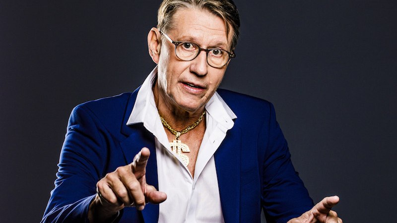 Ein Mann in einem blauen Anzug mit offenem Hemd und goldener Kette gestikuliert vor einem dunklen Hintergrund., &copy; Konzertkreis Triangel Compagnia Compostelli e.V.