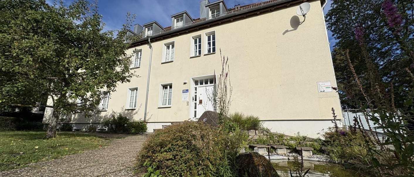 Ein zweistöckiges Gebäude mit hellem Anstrich und mehreren Fenstern. Ein kleiner Garten mit Bäumen und Sträuchern ist im Vordergrund zu sehen., © TOMAS Ein zweistöckiges Gebäude mit hellem Anstrich und mehreren Fenstern. Ein kleiner Garten mit Bäumen und Sträuchern ist im Vordergrund zu sehen., © TOMAS