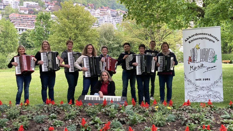 Das Akkordeonorchester Kleinengstingen posiert mit Akkordeons in einem bl&uuml;henden Park. Ein Banner mit dem Orchesterlogo ist sichtbar., &copy; AKK Kleinengstingen
