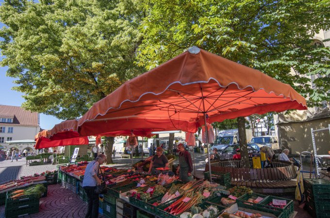 Ein Marktstand mit rotem Sonnenschirm bietet frisches Gem&uuml;se an. Menschen kaufen ein, w&auml;hrend B&auml;ume Schatten spenden., &copy; Stadt N&uuml;rtingen