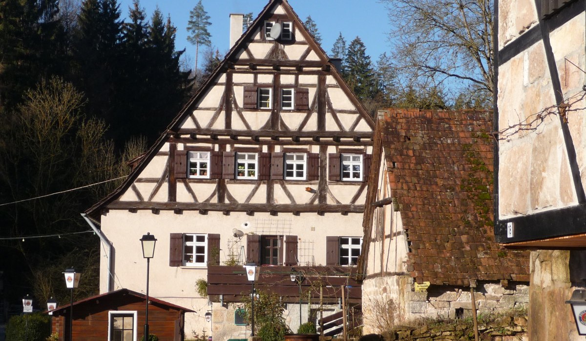 Ein traditionelles Fachwerkhaus mit Satteldach und Holzl&auml;den, umgeben von B&auml;umen und weiteren Geb&auml;uden in einer l&auml;ndlichen Umgebung.