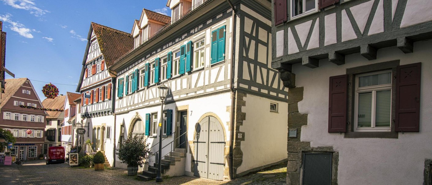 Fachwerkhäuser mit blauen Fensterläden in einer sonnigen Altstadtstraße, dekoriert mit Blumen. Kopfsteinpflaster und klare Himmel im Hintergrund., © Stuttgart-Marketing GmbH, Sarah Schmid Fachwerkhäuser mit blauen Fensterläden in einer sonnigen Altstadtstraße, dekoriert mit Blumen. Kopfsteinpflaster und klare Himmel im Hintergrund., © Stuttgart-Marketing GmbH, Sarah Schmid