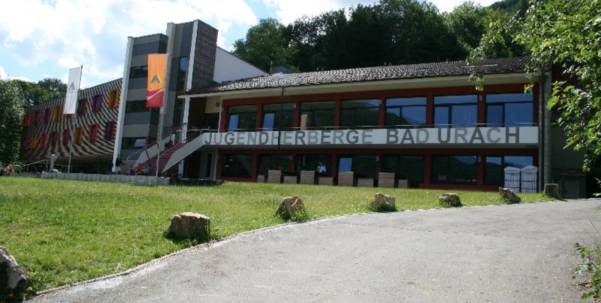 Die Jugendherberge Bad Urach mit moderner Architektur, bunten Fenstern und einem großen Schriftzug. Vorne ein gepflegter Rasen und ein Weg.