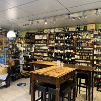 Feinkostladen mit Regalen voller Wein, Spirituosen und Delikatessen. Ein Holztisch mit Stühlen steht im Vordergrund. Der Raum ist hell beleuchtet., © Feinkost-Galeria