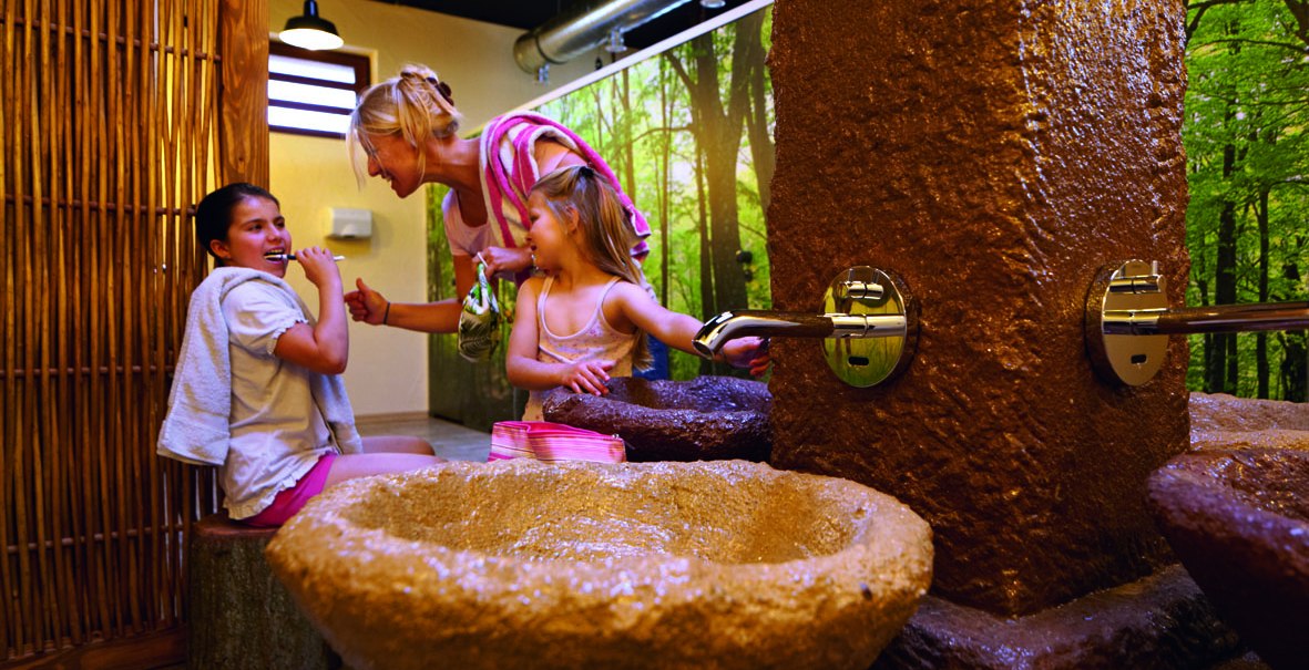 Drei Personen in einem Badehaus mit Naturdekor. Eine Frau und zwei Kinder interagieren fröhlich neben einem steinernen Waschbecken., © Tripsdrill Natur-Resort GmbH